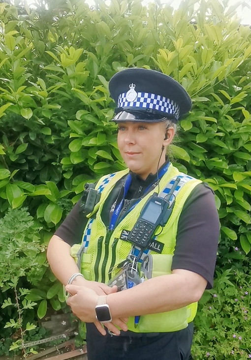 a PCSO in front of a hedge in yellow reflective uniform and police hat
